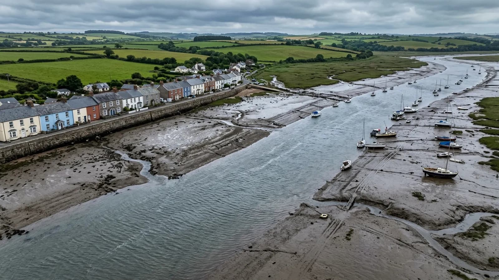 A Devon estuary at half tide from the air, exposed mudflats and channels with boats sitting at angles, a seawall and painted terraces along the shore