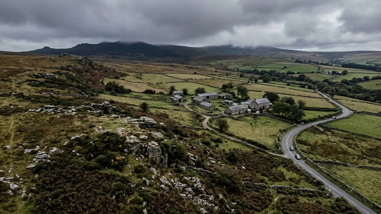 The edge of Dartmoor from the air on a grey day, rough moorland and granite outcrops dropping to walled fields and stone farm buildings with cloud on the high ground