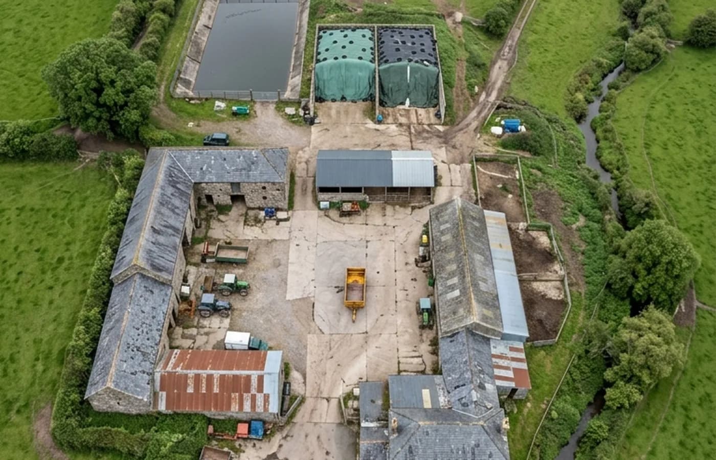 Vertical aerial view straight down over a Devon farmyard from eighty metres, showing stone barns with corrugated and slate roofs around a concrete yard, silage clamps and tracks running out between hedged fields